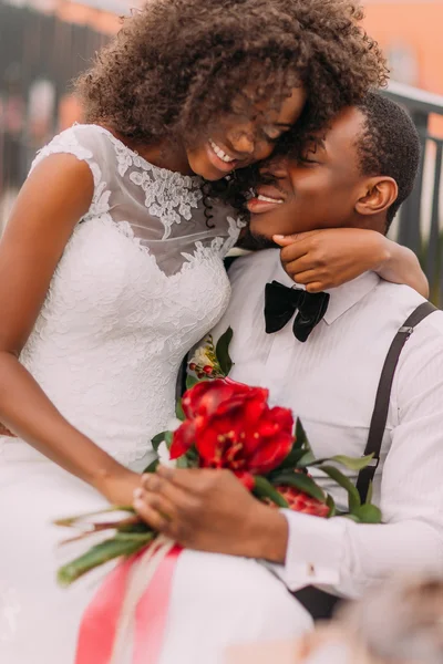 Casal de casamento africano carinhosamente abraçando com buquê de fluxos vermelhos — Fotografia de Stock