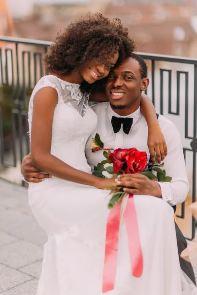 Happy black bride sitting on grooms knees and smiling to camera