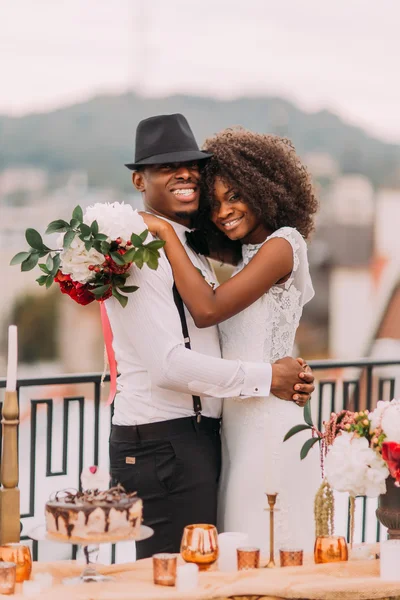 Happy black bride and stylish groom posing for camera on the terrace with cityscape on foreground