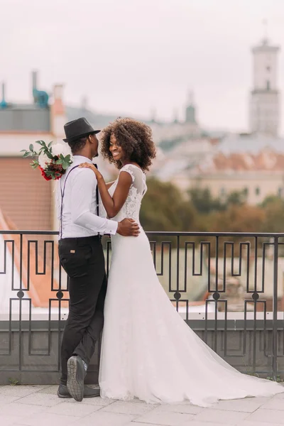 Rozkošní afričtí novomanželé, kteří se navzájem tiše drží a čve. Střešní. Starověká Lvova architektura na pozadí — Stock fotografie