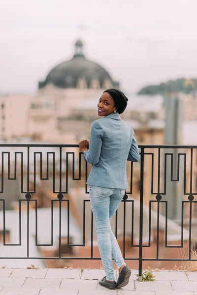 Preciosa chica negra elegante sonriendo a la cámara en la terraza con antigua arquitectura europea en el fondo — Foto de Stock