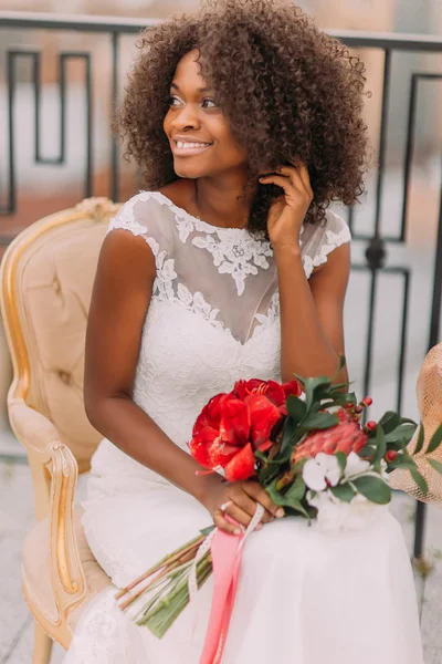 Очаровательная африканская невеста сидит на винтажном терракотовом стуле и счастливо улыбается — стоковое фото