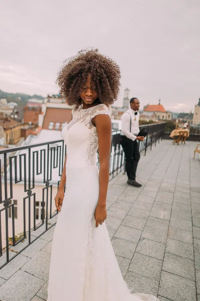 Noiva preta encantadora em vestido de renda vintage no telhado. Noivo elegante confiante no fundo — Fotografia de Stock