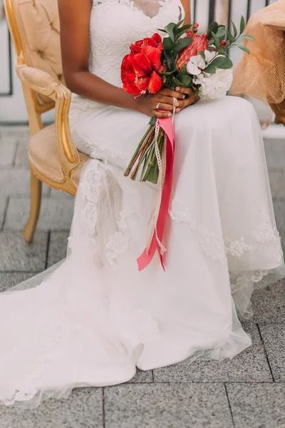Hermosa novia africana sentada en silla con ramo de flores rojas en media longitud — Foto de Stock