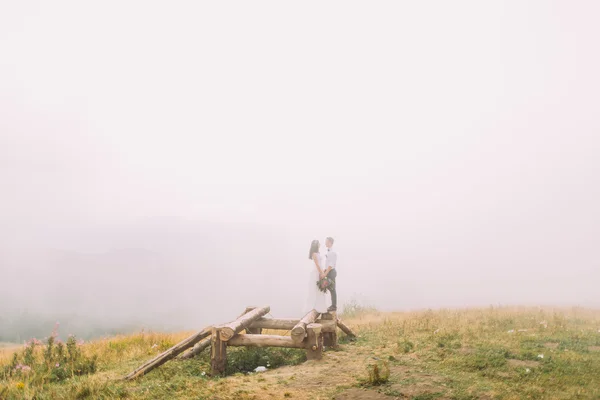 Jonge bruidspaar permanent op de houten brug. Mistige bergen achtergrond — Stockfoto