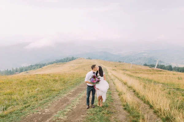 Casamento casal suavemente beijando no caminho nas montanhas nebulosas — Fotografia de Stock