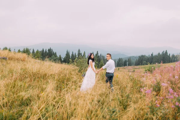 Feliz pareja de boda caminando y tomados de la mano en el campo. Luna de miel en las montañas — Foto de Stock
