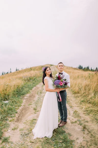 Hermosa pareja de boda con flores de pie en el camino en las montañas —  Fotos de Stock