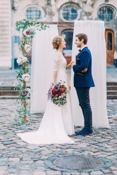 Счастливые молодожены улыбаются и держатся за руки возле свадебной арки, украшенной цветами — стоковое фото