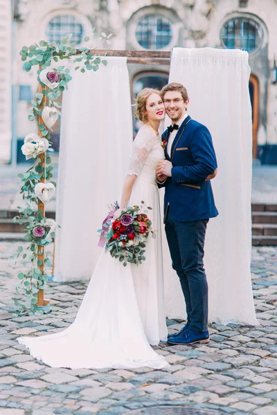 Nádherný pár, usmíval se a pózuje pro fotoaparát poblíž svatební oblouk s květy — Stock fotografie