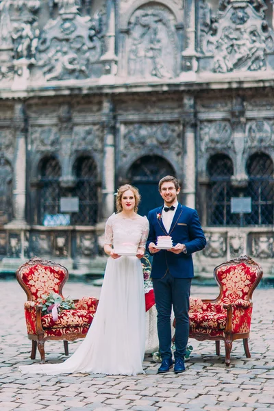 Recém-casados encantadores segurando pedaços de seu bolo de casamento e sorrindo. Arquitetura europeia antiga em segundo plano . — Fotografia de Stock