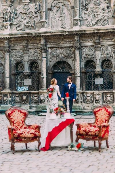 Feliz recém-casados de mãos dadas e amorosamente olhando um para o outro com mesa vintage de luxo e poltronas vermelhas em primeiro plano . — Fotografia de Stock