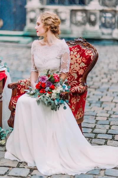 Blonde aristokratische Braut mit Brautstrauß in den Händen auf rotem Vintage-Sessel sitzend — Stockfoto