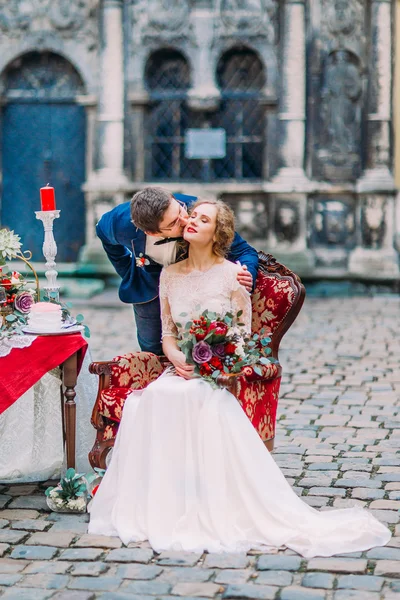 Novio besa suavemente a su aristocrática novia rubia sentada en un sillón rojo vintage —  Fotos de Stock