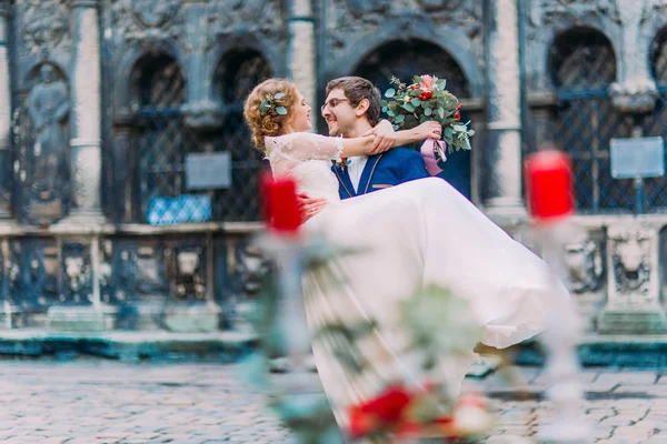 Όμορφος groom κρατά στα χέρια την αριστοκρατική ξανθιά νύφη με το ρομαντικό τραπέζι διακοσμημένο με λουλούδια και κεριά — Φωτογραφία Αρχείου