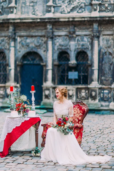 Noiva encantadora com buquê de casamento em mãos sentadas na poltrona vintage e esperando seu noivo com arquitetura europeia velha no fundo — Fotografia de Stock