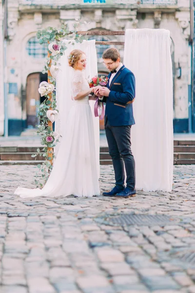 Novia feliz y novio sonriendo y tomados de la mano cerca del arco de la boda decorado con flores —  Fotos de Stock