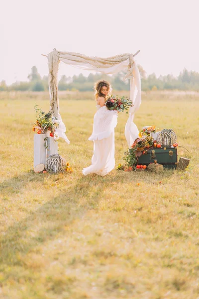 Noiva encantadora em pé no campo com arco de casamento no fundo — Fotografia de Stock