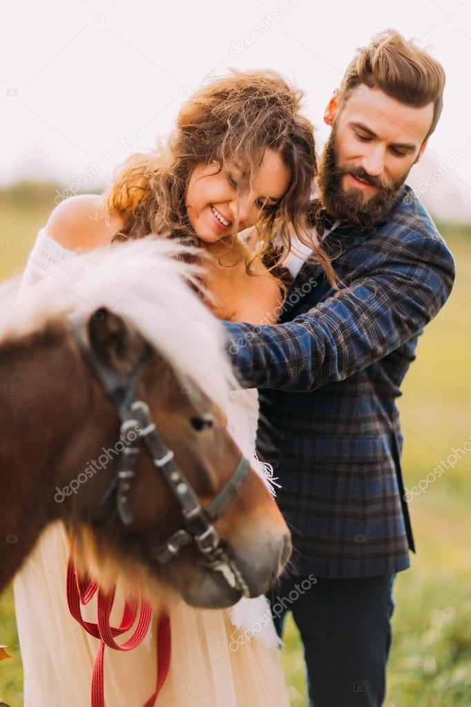 Charming bride and bearded groom stroking pony closeup
