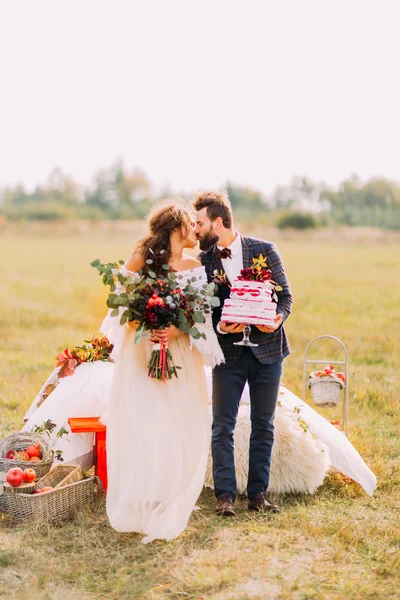 Stijlvolle bruidspaar kussen met boeket en cake in handen — Stockfoto
