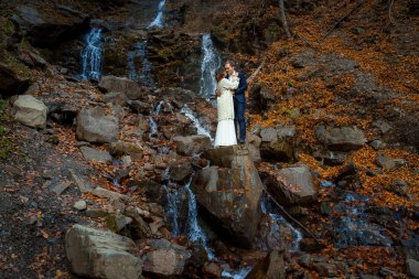 Charming bride and groom stanging on the waterfall. Beautiful autumn forest background