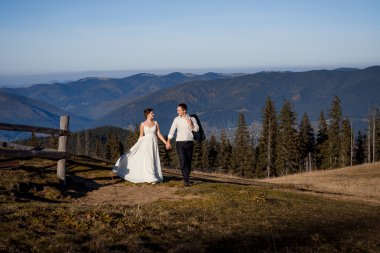 Düğün çifti kırsalkesimde yürüyor ve el ele tutuşuyor. Dağlarda güneşli bir gün