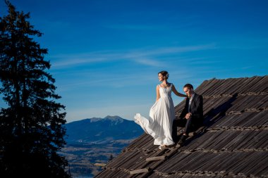 Charming smiling bride and groom standing on the roof of country house. Beautiful mountain landscape background