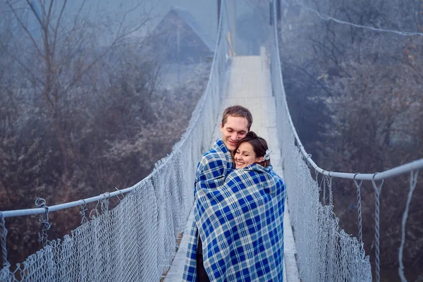 Preciosos novios arrugados en manta abrazos suavemente en el puente de madera. Luna de miel en las montañas — Foto de Stock