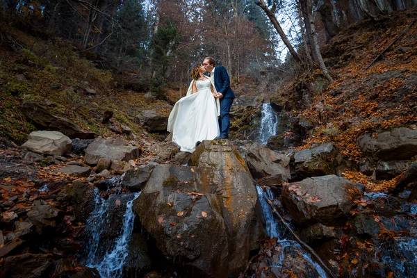 Hermosos recién casados besándose en la cascada. Luna de miel en las montañas de otoño — Foto de Stock