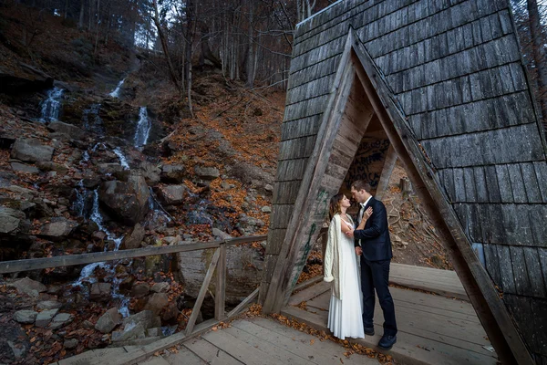 Όμορφο ζευγάρι γαμήλιες αγκαλιές στην ξύλινη γέφυρα στα βουνά. Φόντο καταρράκτη — Φωτογραφία Αρχείου