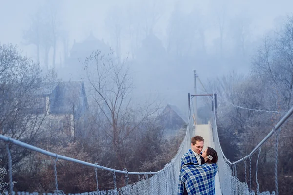 Όμορφο ζευγάρι γάμο τυλιγμένο σε κουβέρτα απαλά αγκαλιές στη γέφυρα ανάρτηση στα βουνά. Φθινόπωρο βουνά φόντο — Φωτογραφία Αρχείου