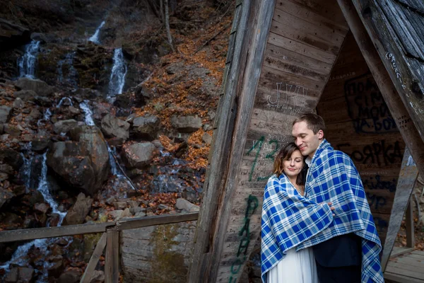 Happy wedding couple wrapped in blanket tenderly hugs with eyes closed. Wodden bridge at the mountains — Zdjęcie stockowe