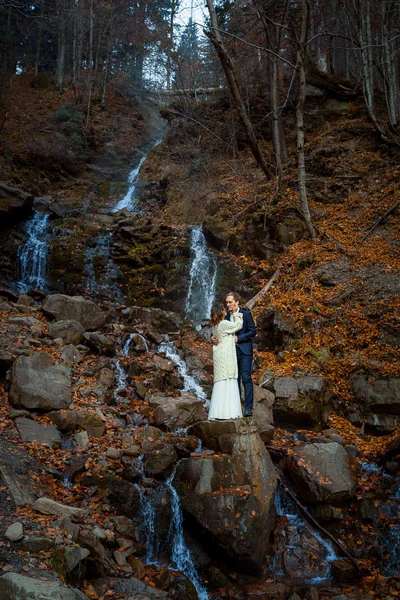 Beautiful wedding couple hugs on the waterfall, Autumn forest background — Zdjęcie stockowe