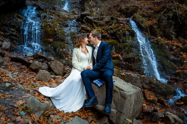 Hochzeitspaar sitzt auf Stein und schaut einander liebevoll an. Hintergrund Wasserfall. — Stockfoto