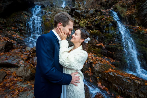 美丽幸福的新娘轻轻地抚摸着她的新郎的面容。在背景上的瀑布。在山中的秋天 — 图库照片