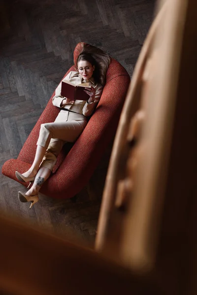 Stylish young girl in beige suit reads a book on the red sofa. Top view