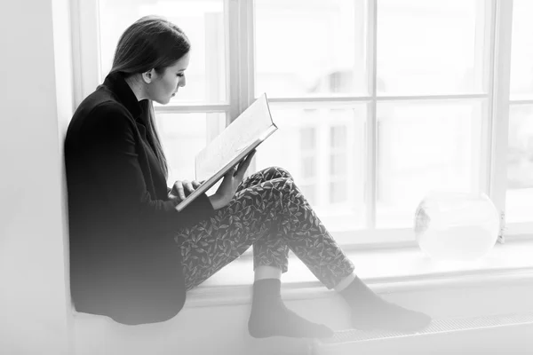Menina bonita e elegante lendo um livro no peitoril da janela. Foto em preto e branco — Fotografia de Stock