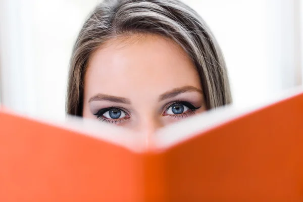 Beautiful blonde student girl covering her face behind the orange book.