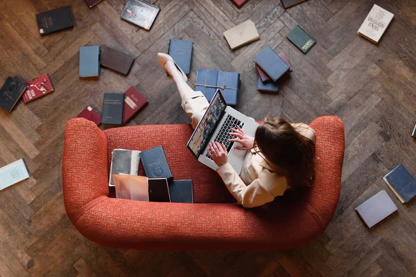 Elegantní mladá dívka v béžové barvy čte knihu na červené pohovce obklopena knihami. Pohled shora — Stock fotografie