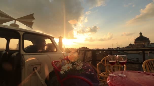 Jovem casal amoroso beijando no carro vintage no café no telhado com copos de vinho tinto e flores na mesa. Vista do antigo edifício da cidade enquanto chovia. Pôr do sol romântico no fundo — Vídeo de Stock