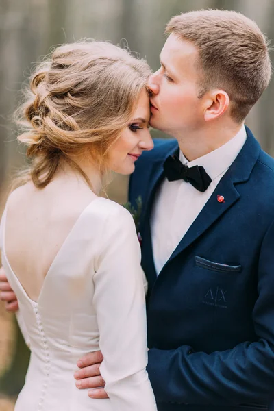 Primer plano retrato de novio con estilo besando a su hermosa novia en la frente. Día soleado en el bosque — Foto de Stock