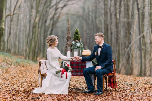 Para ślub siedzi i trzymając się za ręce na świątecznym stole w jesień las — Zdjęcie stockowe