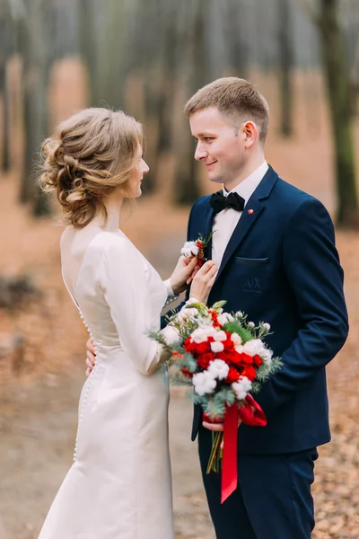 Beautiful bride adjusts boutonniere on the suit of her groom in autumn forest — Stock Photo, Image