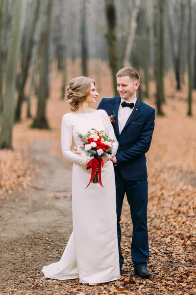 El novio guapo y la novia hermosa se miran amorosamente en el bosque de otoño. — Foto de Stock