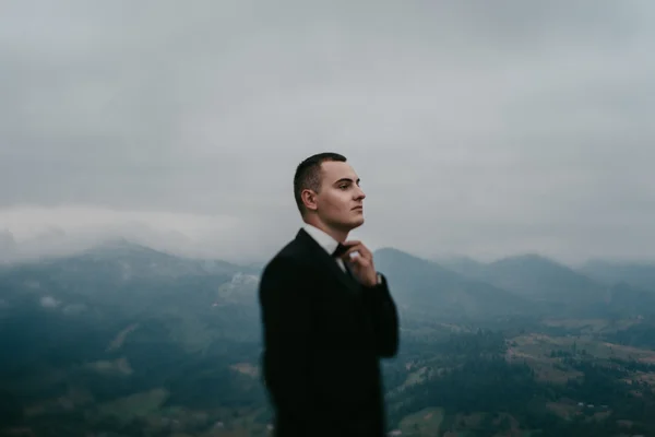 Bonito noivo jovem em pé na frente das montanhas e olhando para a natureza — Fotografia de Stock