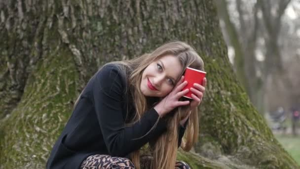 Jonge stijlvolle blonde vrouw die koffie drinkt om in het herfstpark te gaan. Bladloze boom op achtergrond — Stockvideo