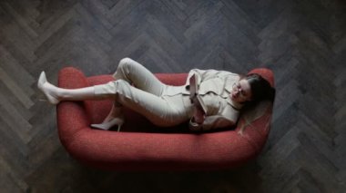 Kitap okurken kırmızı kanepede yatan gözlük ve bej takım elbise güzel sarışın genç kadın. Kız gözlük ayarlama ve eğlenceli kameraya bakıyor. Üstte görüntü