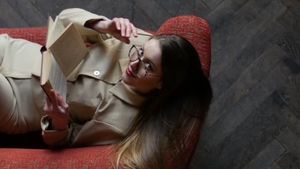 Bella bionda giovane donna in occhiali e abito beige seduta sul divano rosso durante la lettura di libri. Ragazza che regola gli occhiali e guarda giocosamente nella fotocamera. Vista dall'alto — Video Stock