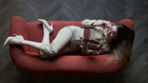 Attraente ragazza bionda tatuata in occhiali e abito beige in posa giocosa e sorridente sul divano rosso durante la lettura del libro. Vista dall'alto — Video Stock