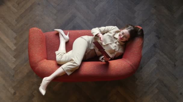 Mujer joven seductora hermosa en gafas y traje beige posando juguetonamente y sonriendo en el sofá rojo mientras lee el libro. Vista superior — Vídeos de Stock